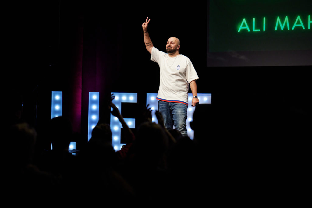 Ali Mahlodji, CEO futureOne steht auf einer Bühne und zeigt das Peacezeichen zum Publikum